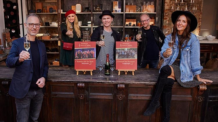 Fünf Personen stehen an einer Bar, halten Sektgläser und lächeln, während sie auf zwei Plakate mit Veranstaltungen zeigen.