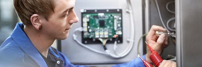 Ein junger Techniker arbeitet konzentriert an einem elektrischen Gerät, während er mit einem Schraubendreher und Messleitungen hantiert.