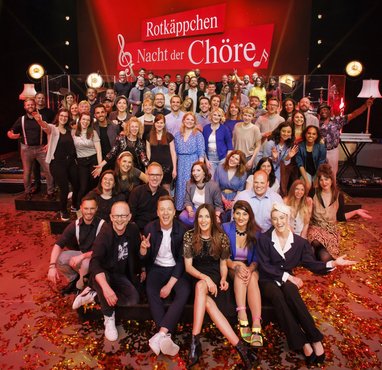 Die Teilnehmer der 'Nacht der Chöre' stehen zusammen, lächeln und zeigen ihre Begeisterung in einem festlichen Ambiente.
