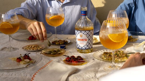 Eine Hand greift nach einer Olive, während zwei Gläser mit orangen Getränken und eine Flasche Canonita im Hintergrund stehen.