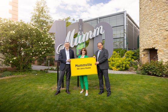 Drei Personen stehen im Freien vor einem großen Mumm-Schild, das 100 Jahre Mumm feiert, und halten ein gelbes Schild mit 'Mummville'.