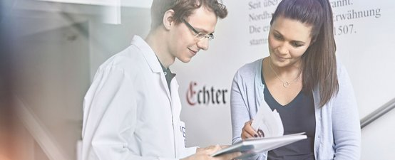 Ein junger Mann im Laborkittel und eine Frau besprechen Dokumente, während sie in einem modernen Raum stehen, beide wirken interessiert.