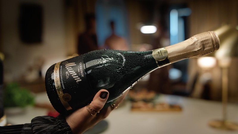 Eine Hand hält eine feuchte Flasche Mumm-Sekt, während im Hintergrund unscharfe Personen und eine gemütliche Atmosphäre zu sehen sind.