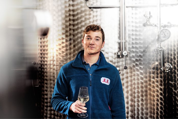 A young man in a blue fleece jacket holds a glass of white wine and smiles in a modern wine production environment.